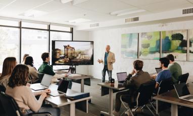 A group of university students seated with laptops in a classroom setting listen to a professor in front of a screen showing ancient ruins.