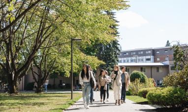 A group of people walk along a pathway in a campus-like setting, surrounded by trees, grass, and buildings.