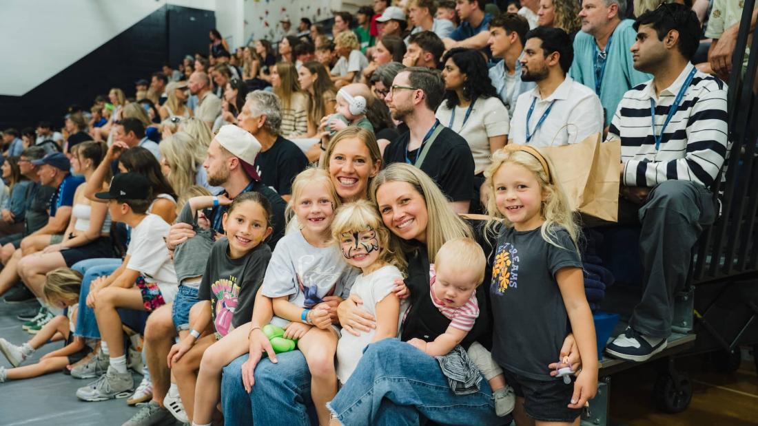Crowd seated in a gym for Homecoming 2025, with two smiling women and children in the foreground.