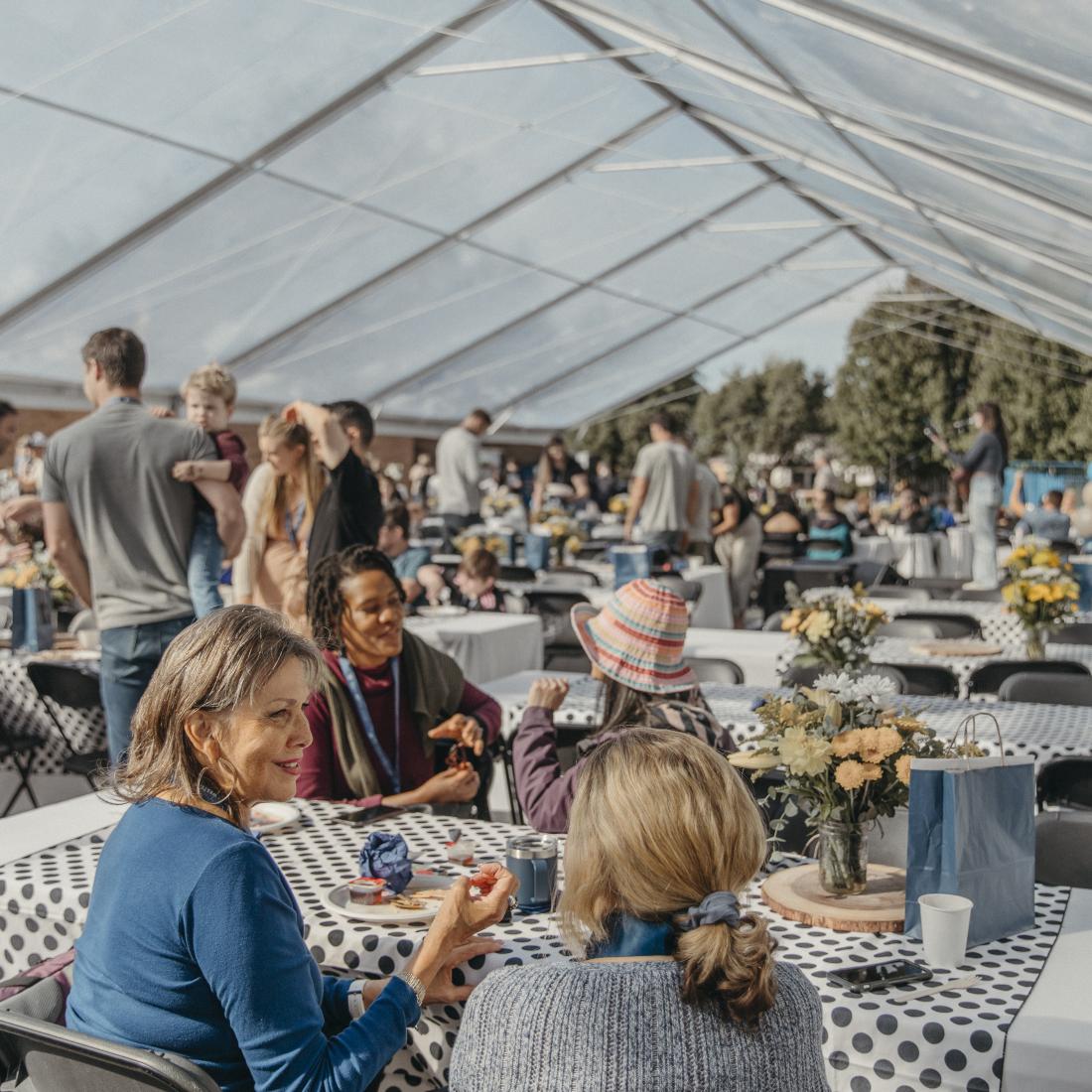 Large group of people gathered under a transparent tent with decorated tables, eating, conversing, and mingling.