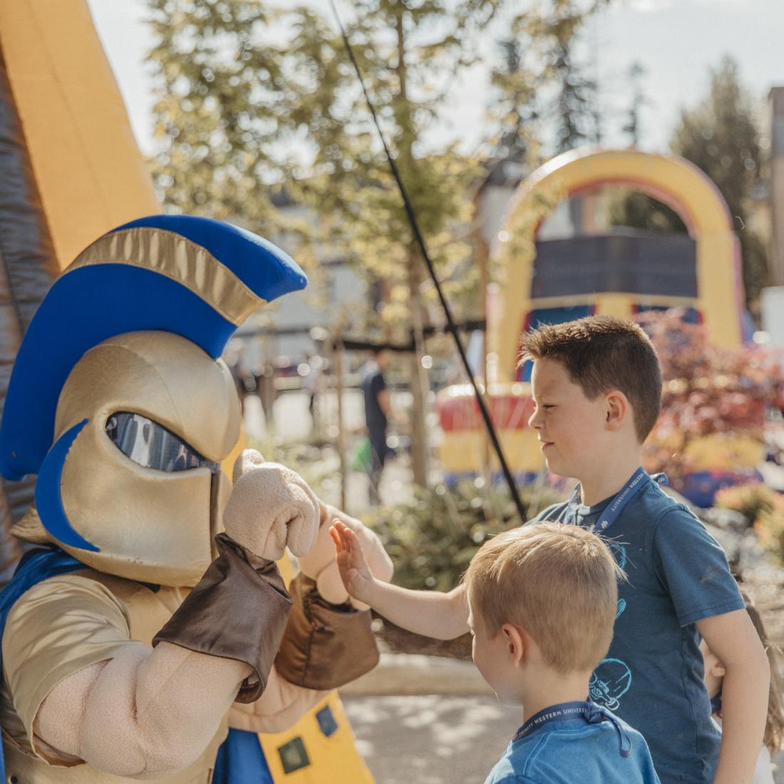 Person in a gladiator costume giving fist bumps to two children outdoors, with an inflatable structure and trees in the background.